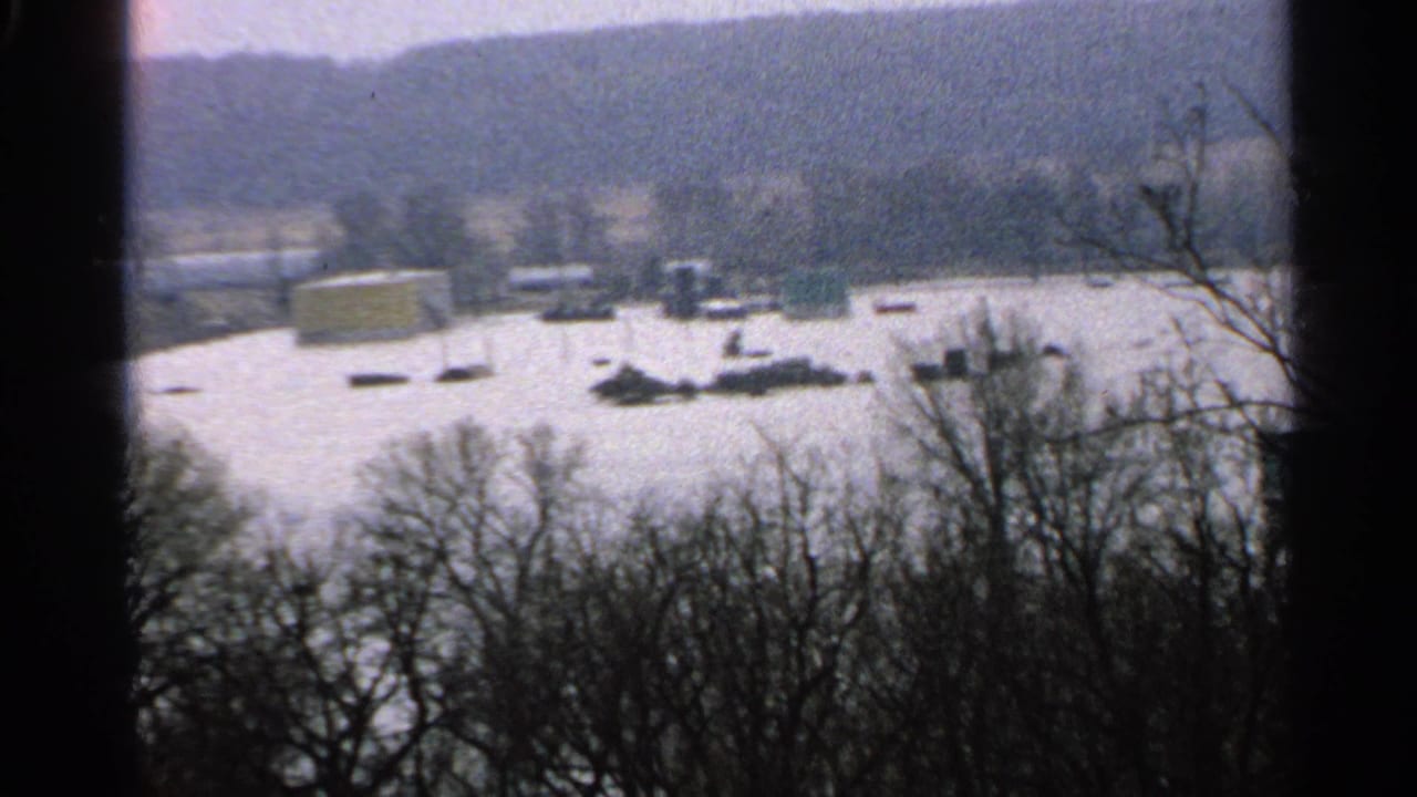Minnesota 1965 8mm frame showing a snow-covered landscape seen through bare trees, with distant structures or objects across the white ground.