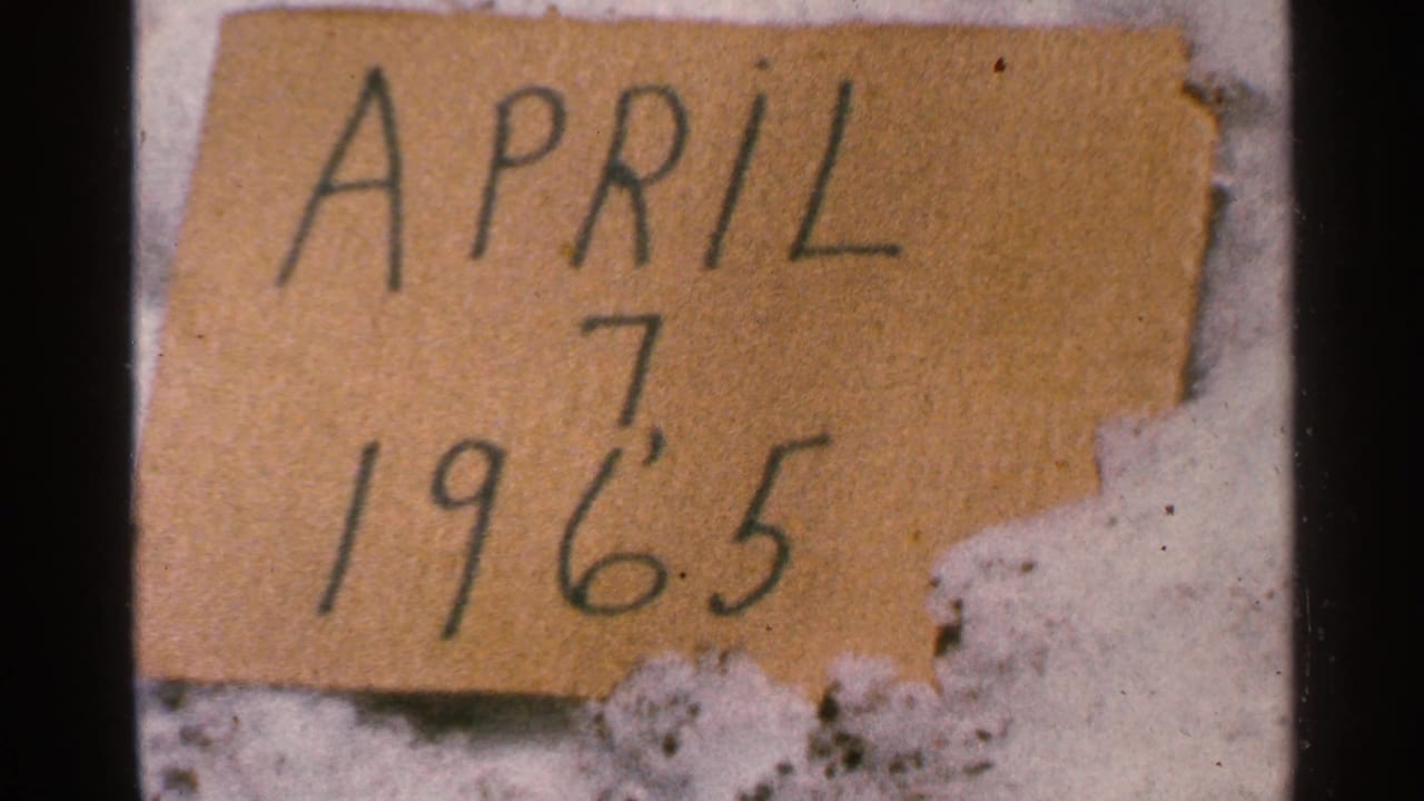 Close view of a worn cardboard sign reading April 7 1965 in a 1965 Minnesota 8mm home-movie frame.