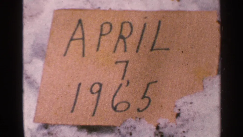 Close 8mm frame of the handwritten April 7 1965 cardboard marker with torn edges and snow behind it.