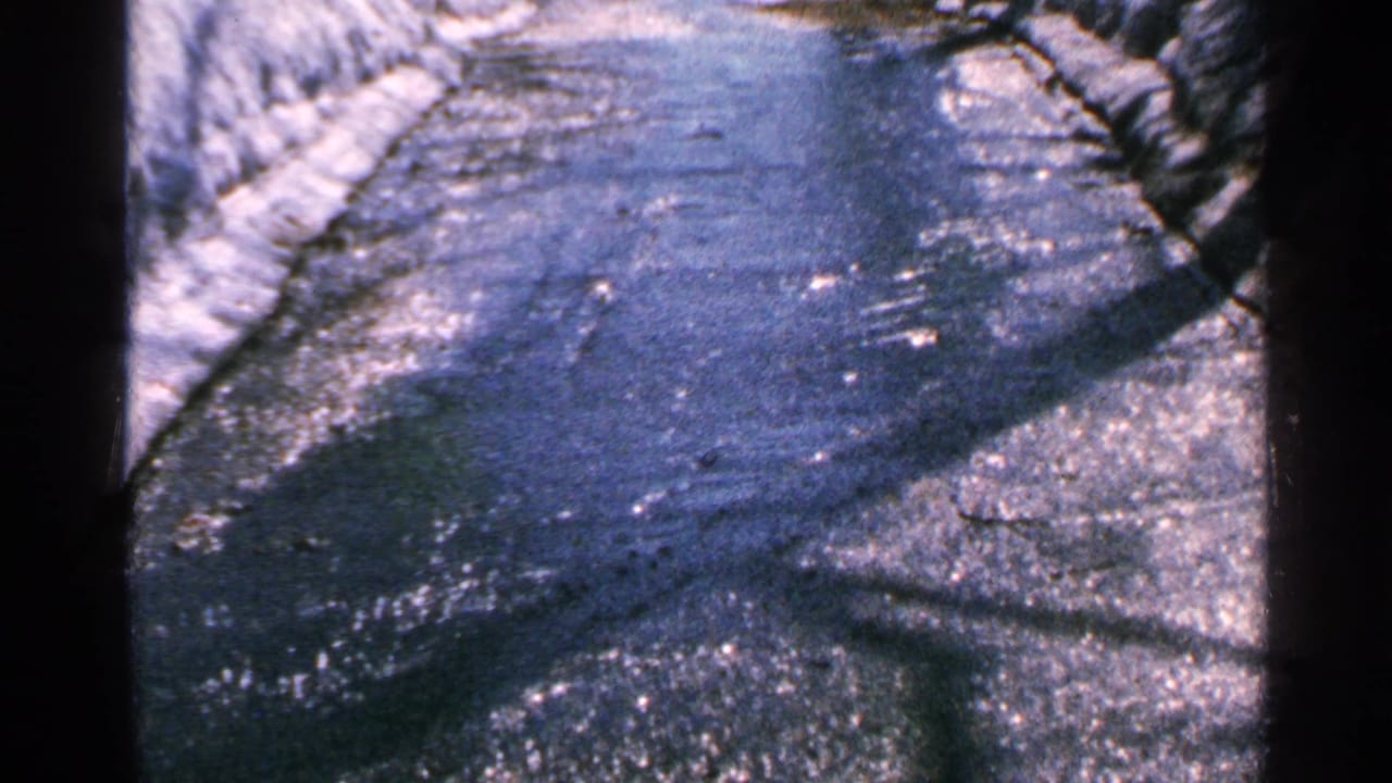 1965 Minnesota 8mm frame showing water flowing over pavement beside snowbanks and dark tree shadows.