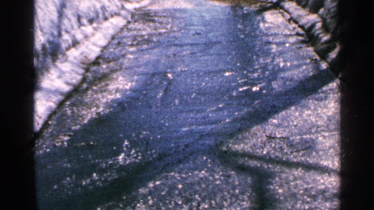 1965 Minnesota home-movie frame of a wet road surface with runoff, snow at the sides, and strong shadow patterns.