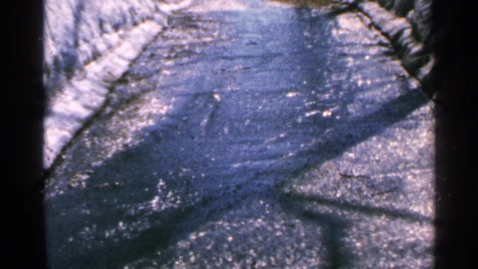 1965 Minnesota home-movie frame of a wet road surface with runoff, snow at the sides, and strong shadow patterns.