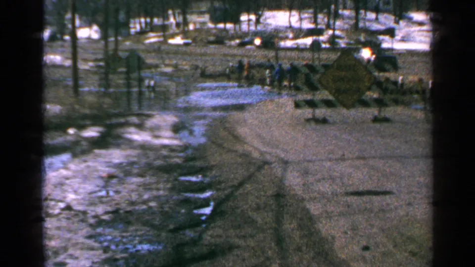 1965 Minnesota 8mm view of a blocked flooded roadway with a yellow warning sign, barricade, snow patches, poles, and distant people.