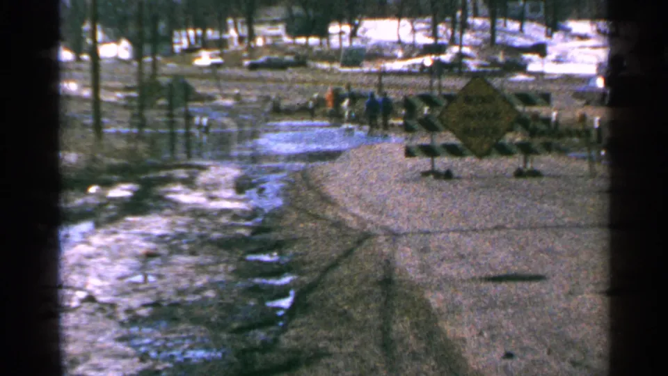 1965 Minnesota home-movie frame showing a flooded road closure with a warning sign, barricade, utility poles, bare trees, and distant bystanders.