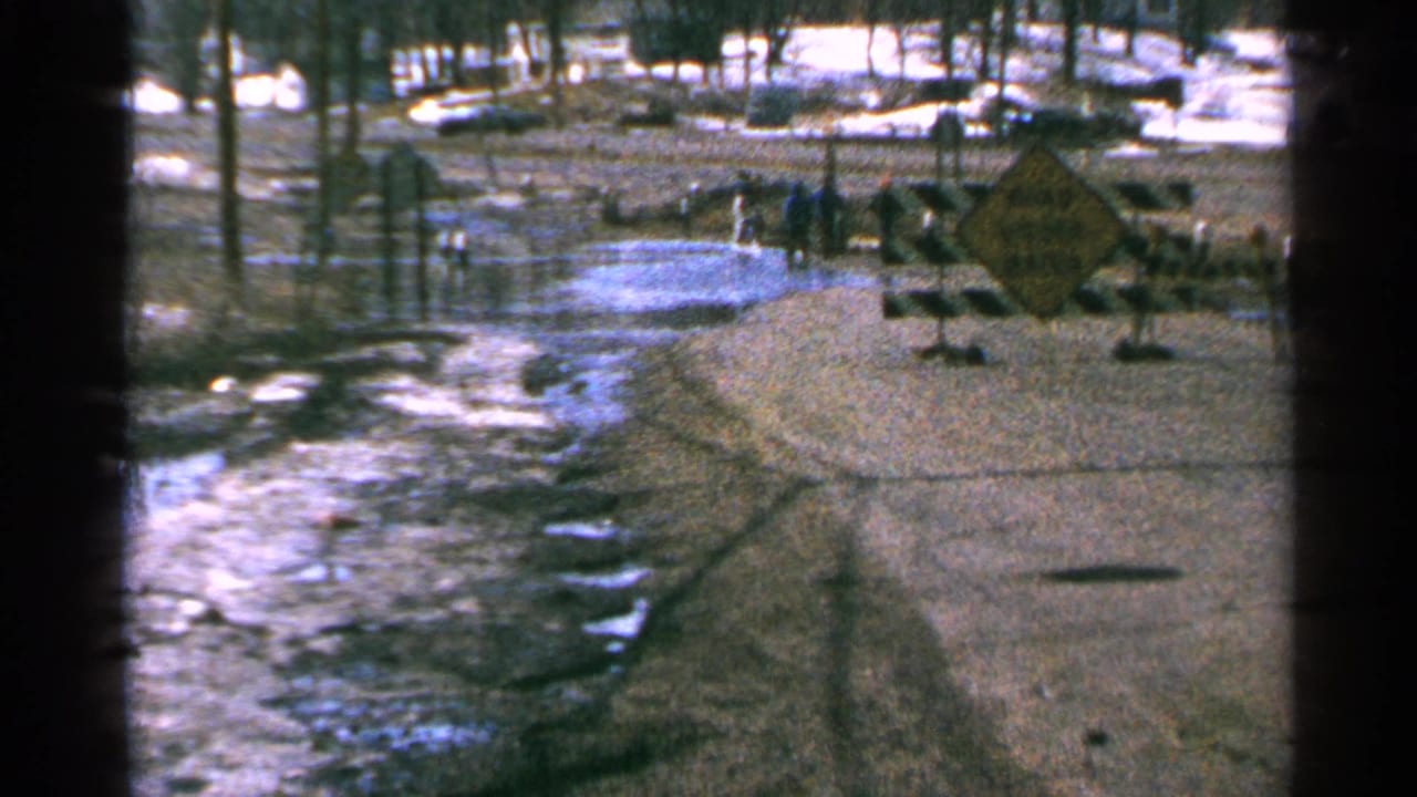 Flooded winter road with snow patches, bare trees, barricades, and people near a warning sign.