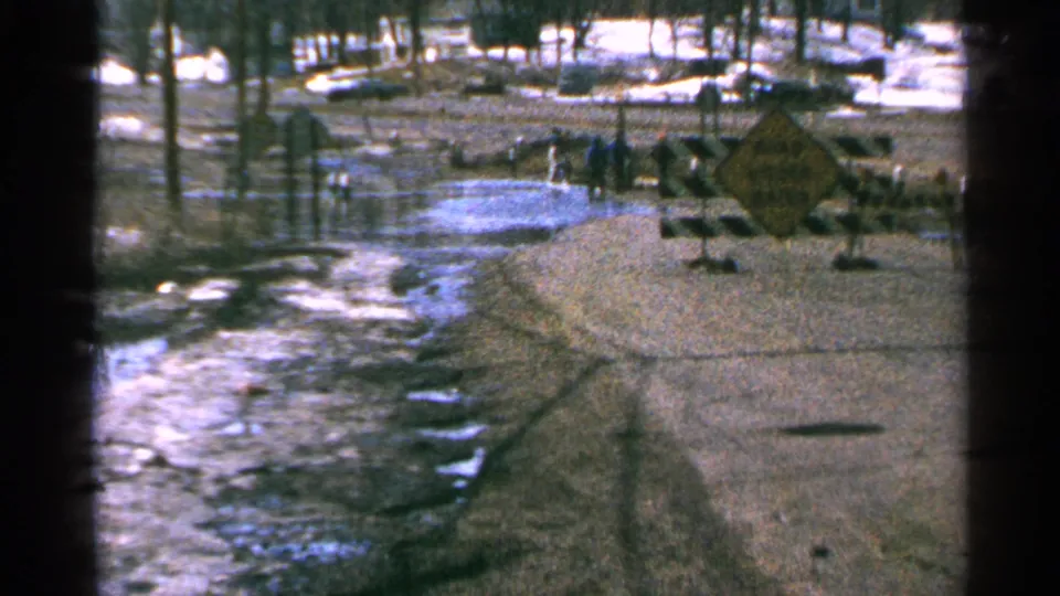 Minnesota, 1965: flooded rural road with icy water, snow patches, bare trees, striped barricades, and bundled people near a yellow warning sign.