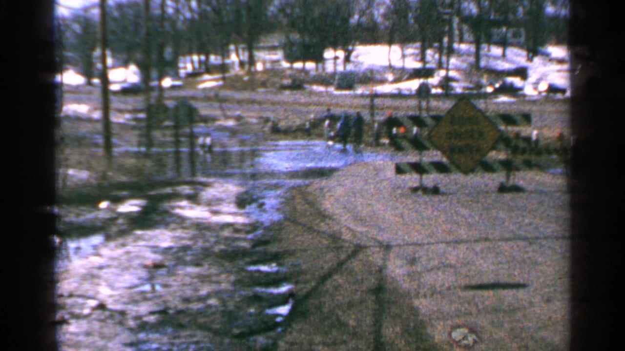 Minnesota, 1965: icy floodwater reaches the edge of a rural road while people stand near striped barricades and a yellow roadside warning sign.