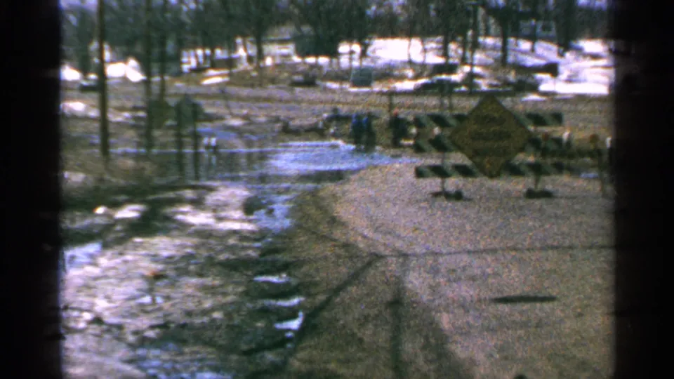 Minnesota, 1965: snowy roadside floodwater reflects bare trees as distant people stand near barricades and a yellow warning sign.