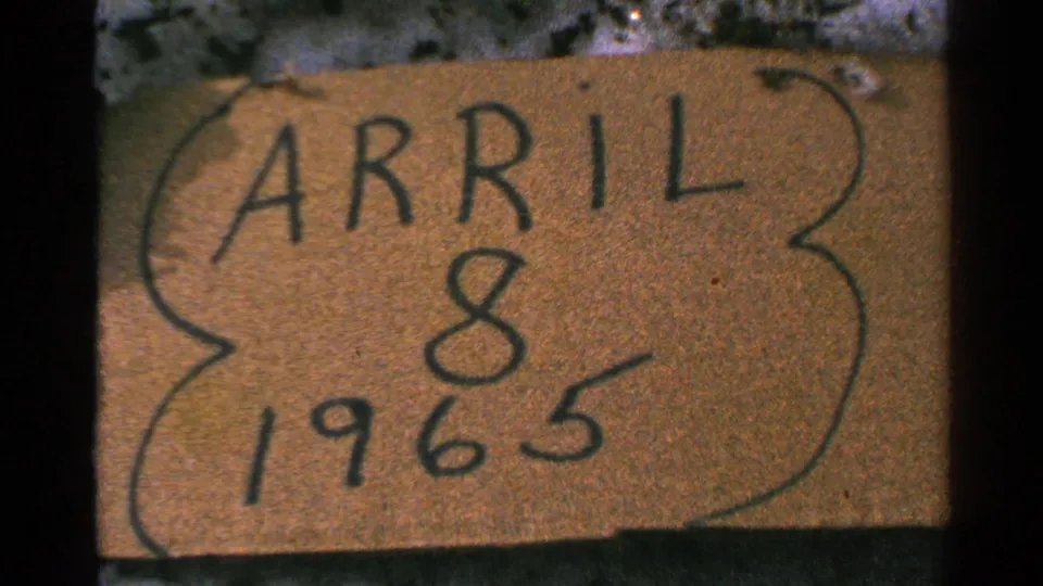Close 8mm home-movie frame of a cardboard date sign reading APRIL 8 1965 in dark marker.