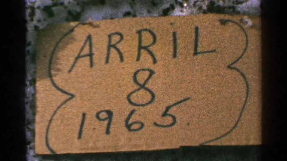1965 home-movie date card fills the frame, with APRIL 8 1965 handwritten on cardboard.