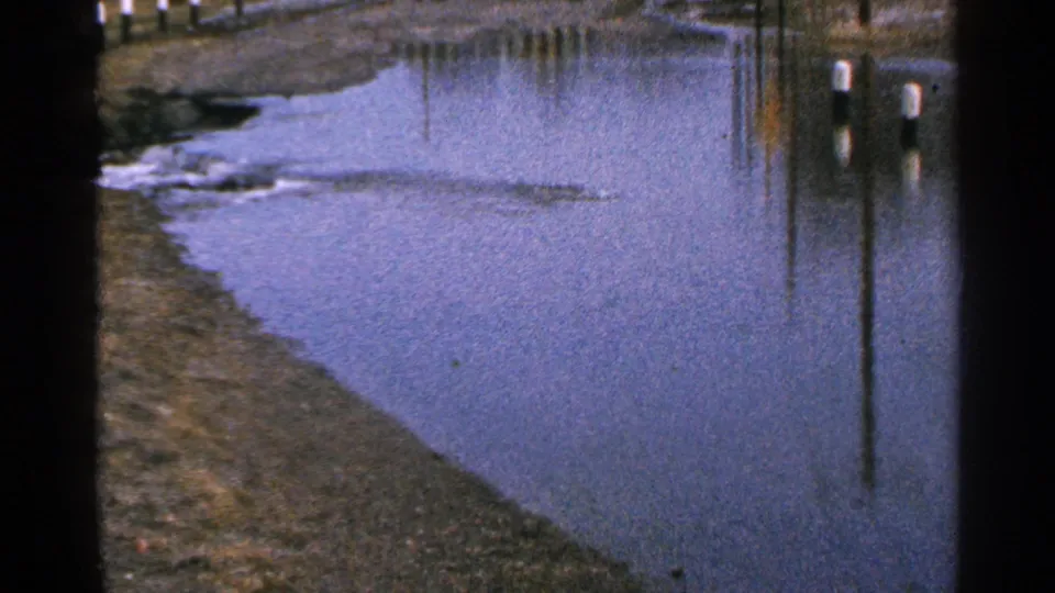 Static 8mm view of standing water beside a roadside in Minnesota, with a sloped muddy edge and vertical pole reflections.