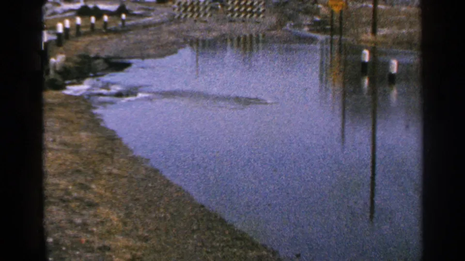 1965 Minnesota home-movie frame showing roadside flooding, vertical reflections, marker posts, trees, and a small yellow sign in the distance.