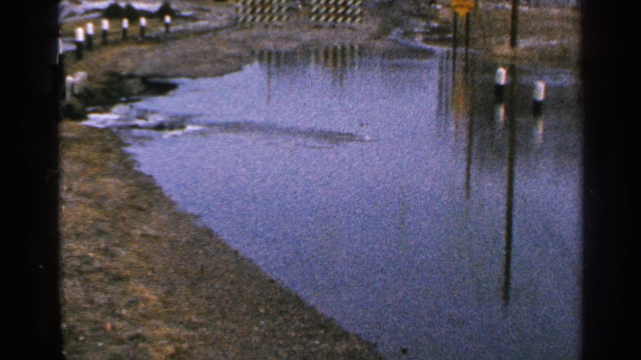 Minnesota 1965 8mm frame showing pooled roadside floodwater, muddy banks, white-topped posts, and a yellow sign reflection.