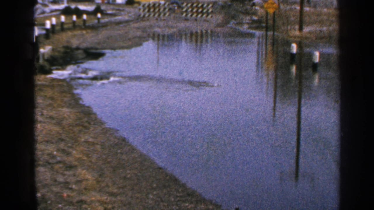 Minnesota 1965 8mm frame of a flooded roadside area with still water, white-topped posts, muddy edges, and a yellow sign.