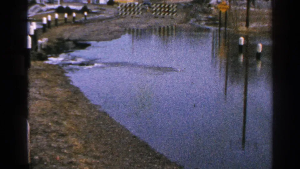 Minnesota 1965 8mm frame showing a broad pool of roadside water, post reflections, muddy banks, and a yellow sign in the distance.