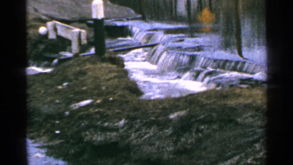 Minnesota 1965 8mm frame showing runoff flowing over a small concrete spillway near roadside posts and wet ground.