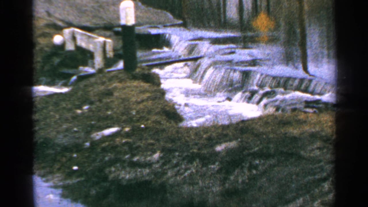 1965 Minnesota 8mm frame showing water spilling over small dam drops beside dark banks, wooden edging, and a white-topped post.