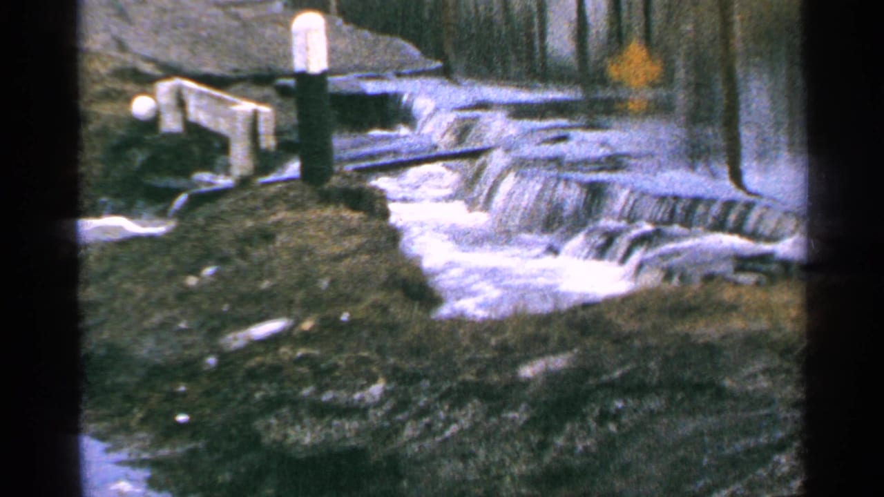 1965 Minnesota 8mm frame of foamy river water crossing a low dam, with a white-topped post and shadowed bank near the edge.