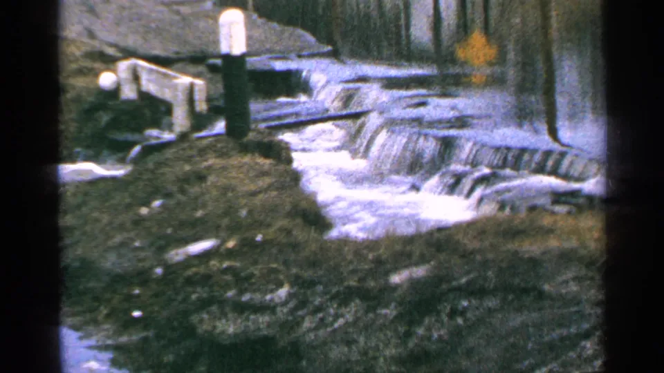 1965 Minnesota 8mm frame of foamy river water crossing a low dam, with a white-topped post and shadowed bank near the edge.