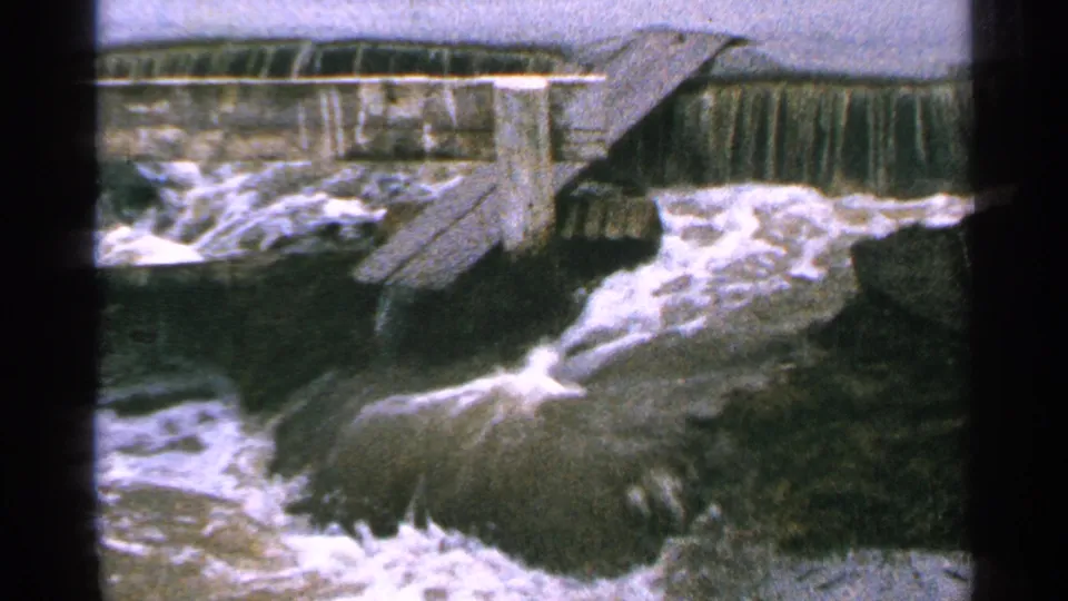 1965 Minnesota 8mm frame showing whitewater below a small wooden dam structure, with dark rocks or banks around the flow.