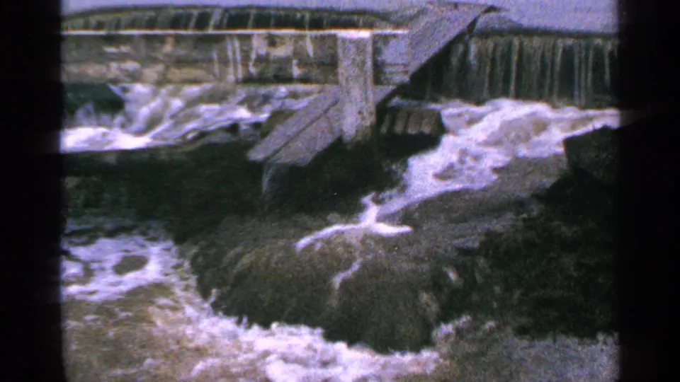 1965 Minnesota 8mm frame of river water spilling through a low dam area, bordered by wooden boards and dark banks.