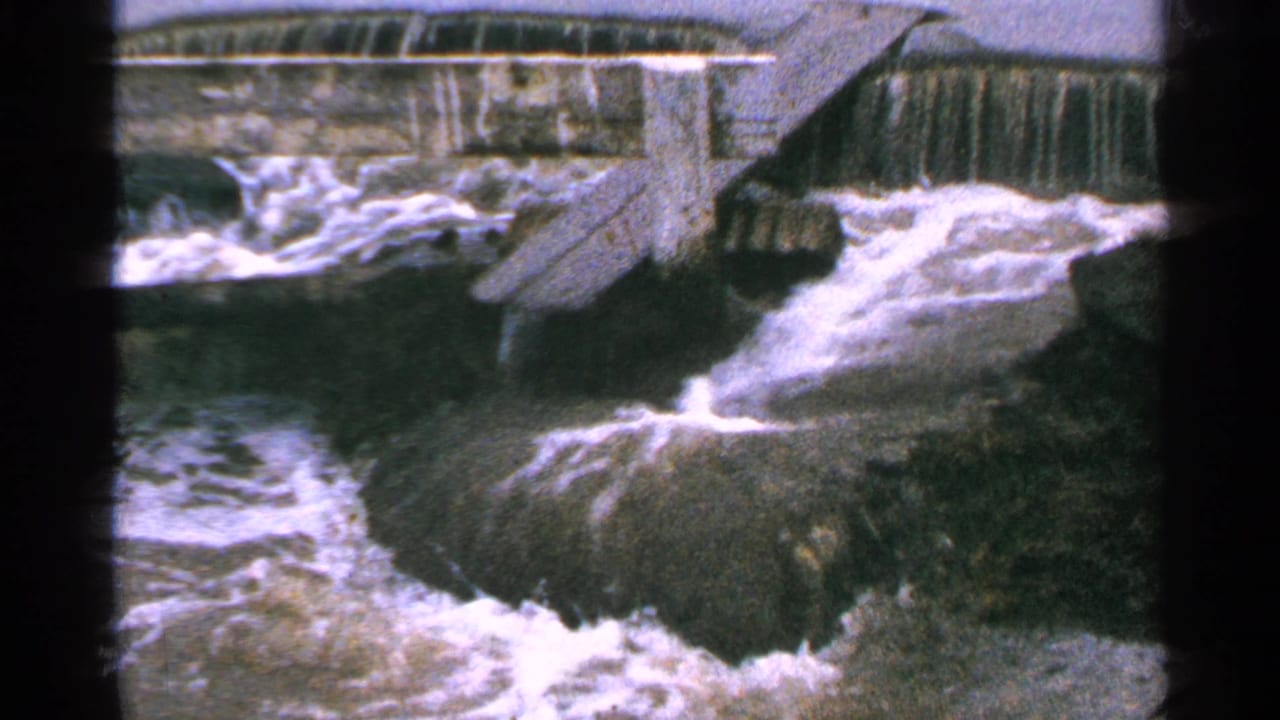 Whitewater surges around rocks beneath a dam spillway.