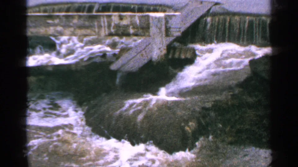 1965 Minnesota home-movie frame with rough water pushing past rocks below a concrete dam.