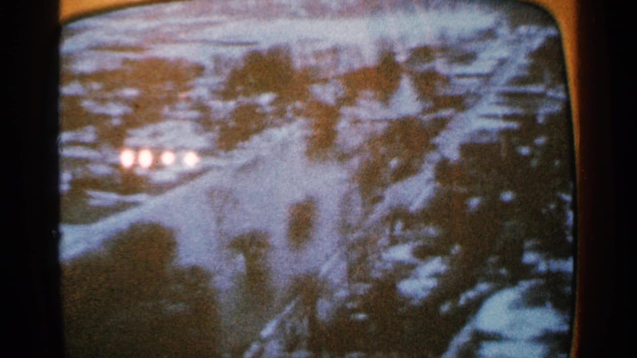 1965 8mm frame from Minnesota metadata showing a grainy rounded-screen view of snow-covered ground, dark shapes, and four bright lights in the distance.
