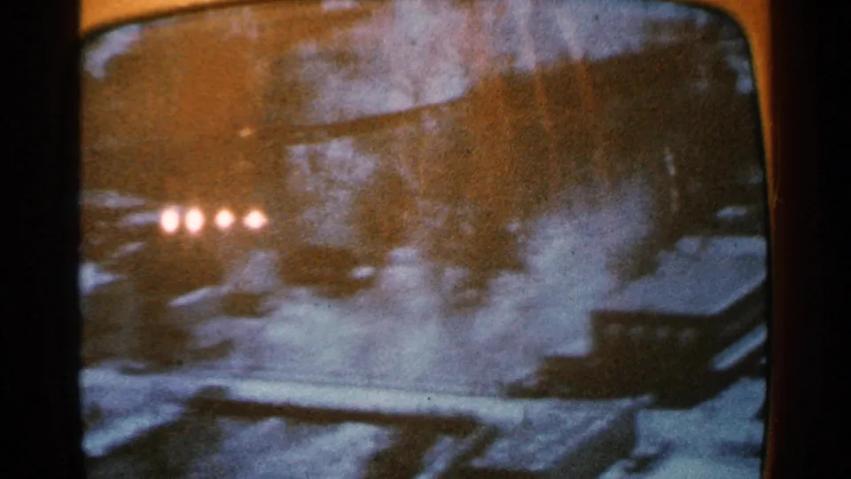 Final 1965 8mm frame showing a grainy rounded-screen snowy landscape with four bright lights, shadowed trees or shrubs, and indistinct terrain.