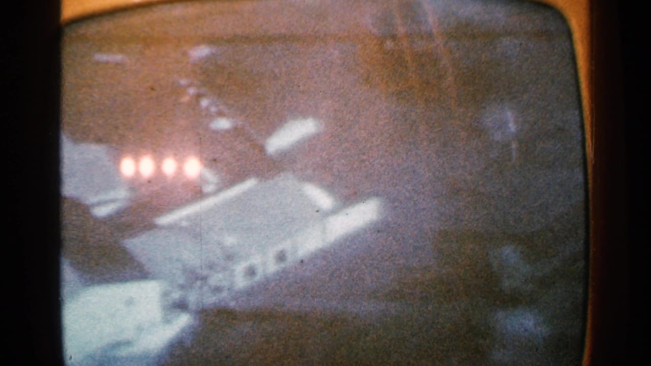 1965 8mm frame from Minnesota showing a grainy snowy view on a rounded screen, with bright lights and a pale angled form.