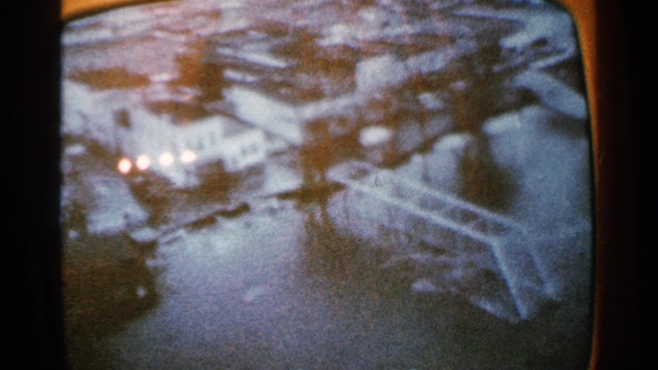 Aerial view of a flooded town with buildings, water, and bright vehicle lights.