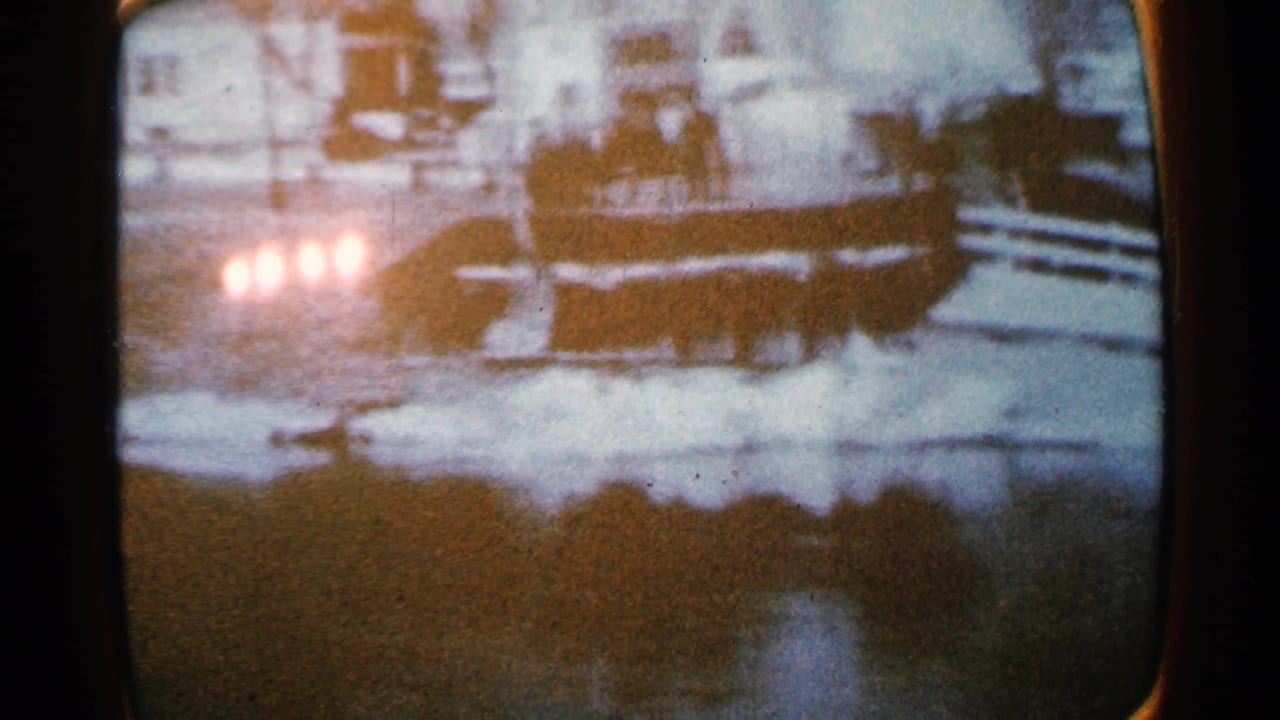 1965 Minnesota 8mm frame showing snow along a partially frozen river or roadside, with soft headlights and a dark vehicle shape in the distance.