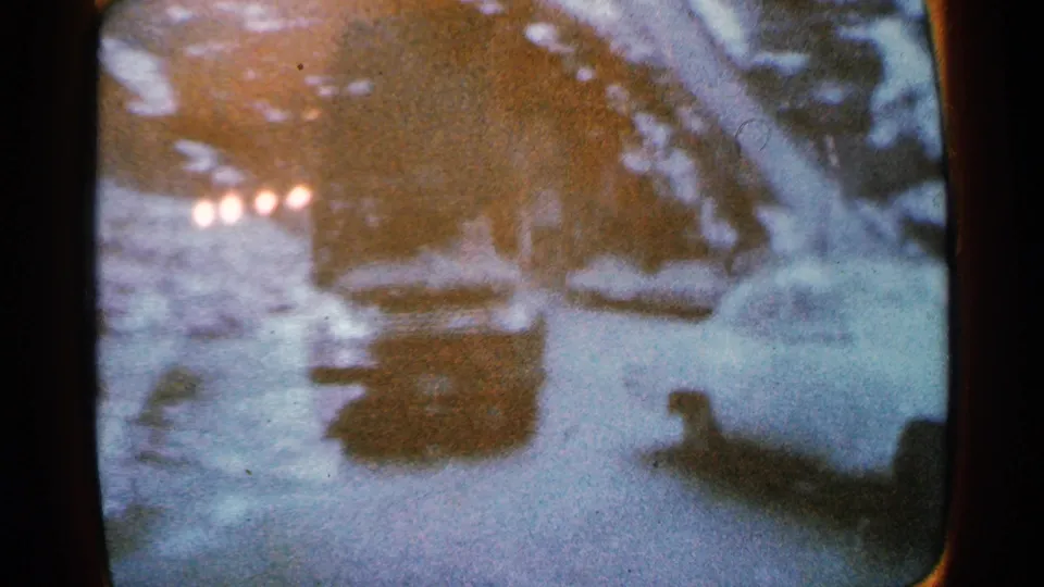 1965 Minnesota winter roadside view beside a partially frozen river, with snow, parked cars, and small glowing headlights in soft focus.