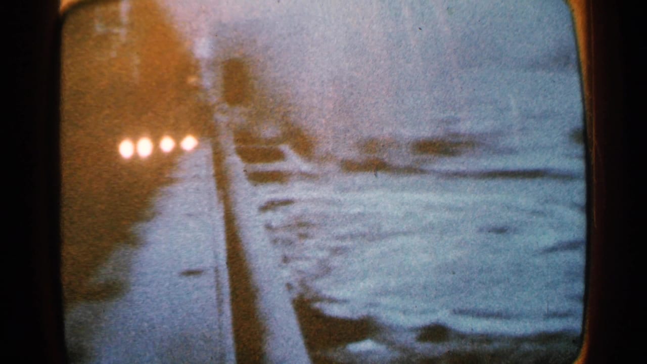 1965 8mm frame of a television broadcast showing a snowy street scene in Minnesota with vehicles and screen glare.