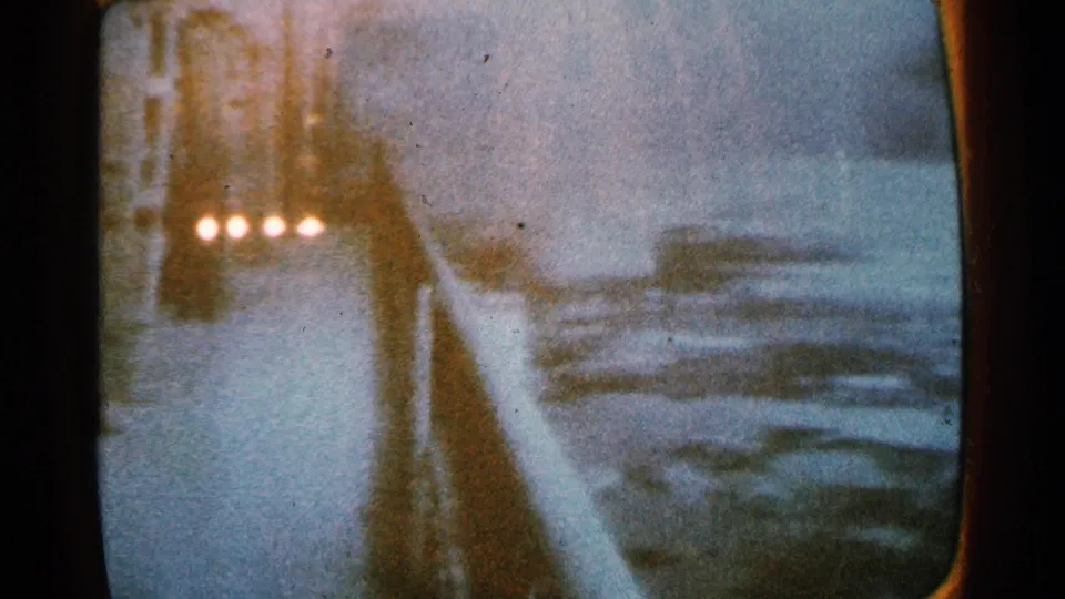 Filmed television broadcast showing a snowy winter road scene with a dark vehicle and soft screen reflections.