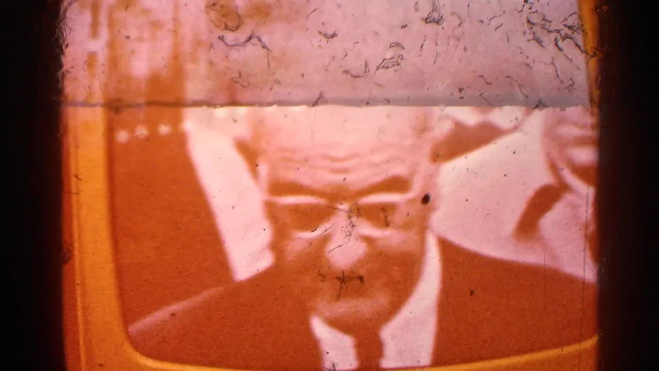 Warm, orange-tinted 8mm frame of a televised close-up showing an older suited man wearing glasses.