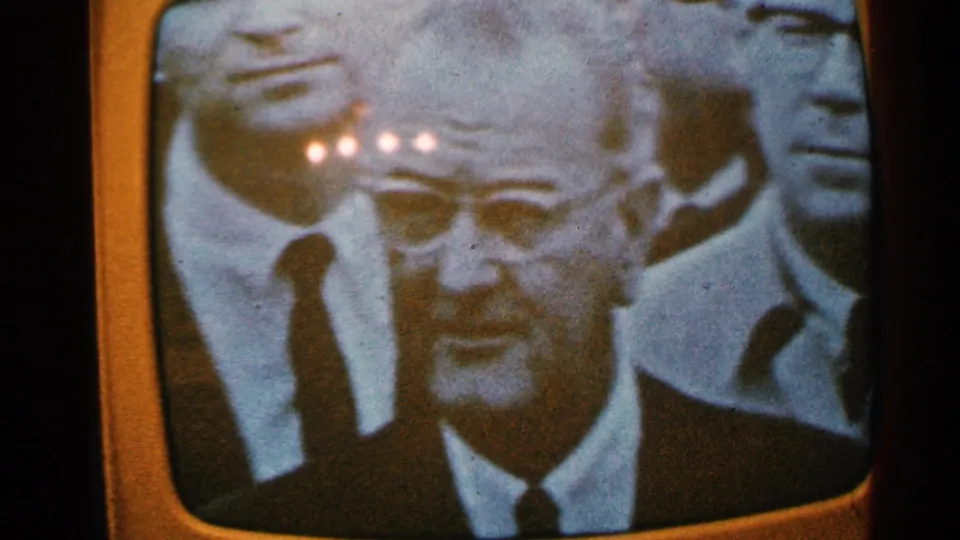 1965 home-movie frame of a television news shot showing a suited man with glasses and partial figures behind him.