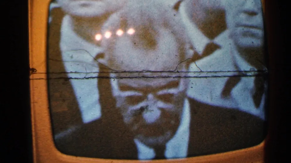 Minnesota 1965 8mm frame of an old television screen showing a black-and-white news broadcast with suited men, reflections, and scan distortion.