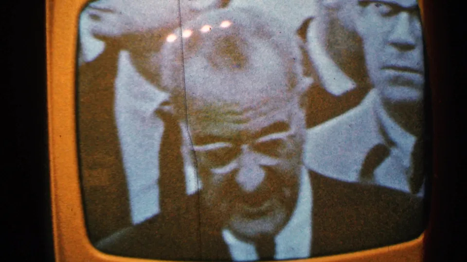 Minnesota 1965 home-movie frame showing a monochrome TV news close-up of a suited man with other faces behind him.