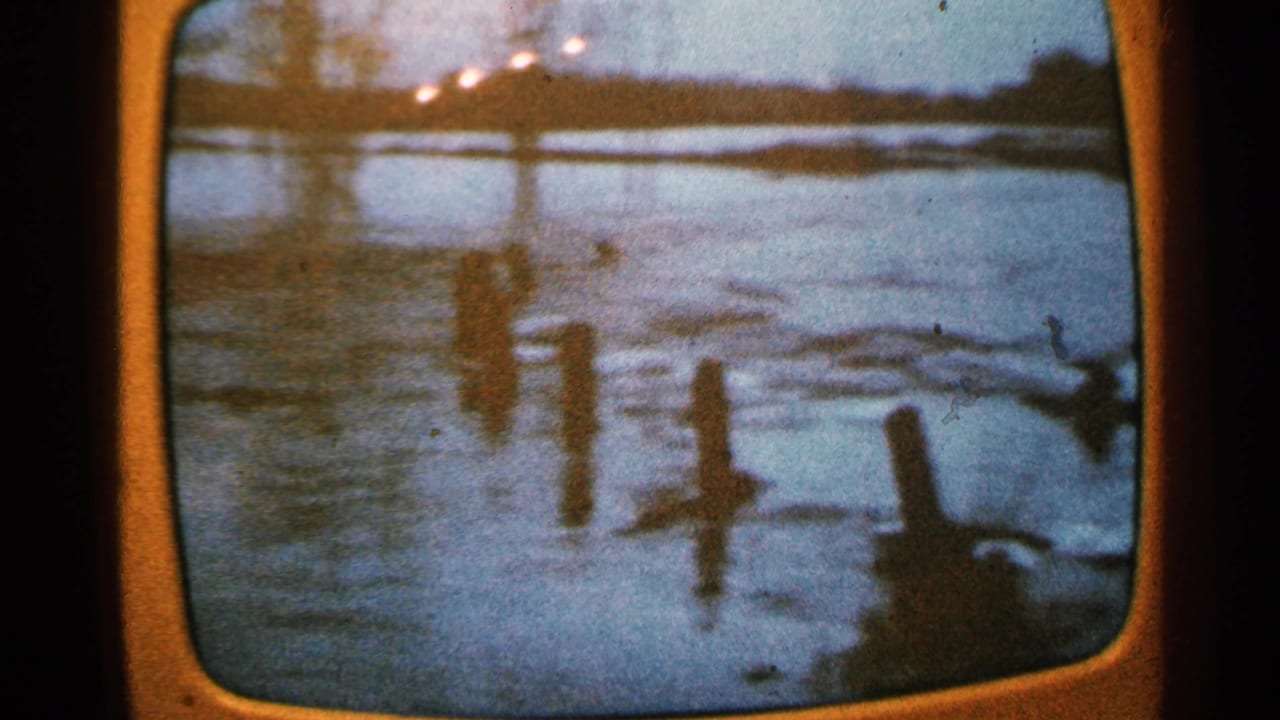 A rounded television screen shows floodwater, wooden posts, and a dim distant shoreline in 1965 Minnesota footage.