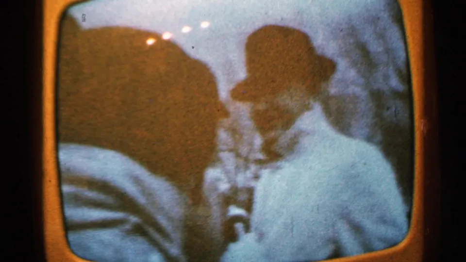 Minnesota 1965 home-movie frame showing a bundled adult near the camera, with soft focus, orange borders, and projected-film reflections.