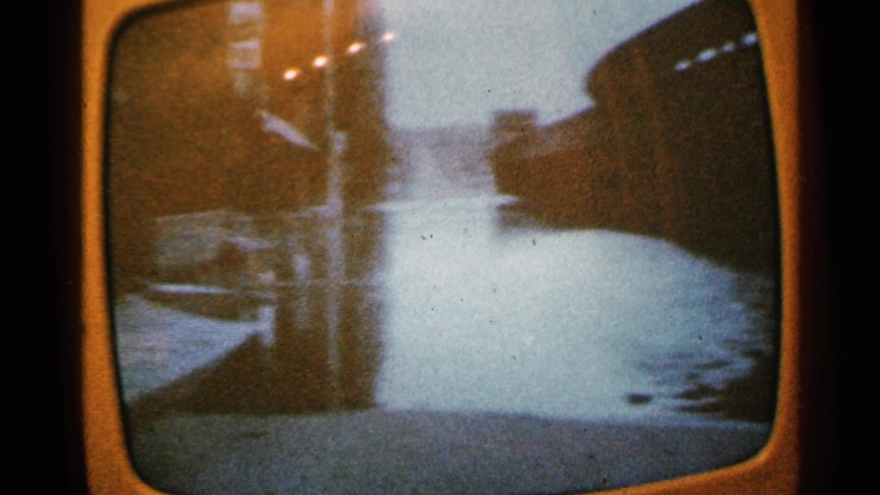 1965 Minnesota 8mm frame of a rounded television screen showing a flooded street or waterfront with dark structures and reflected light.
