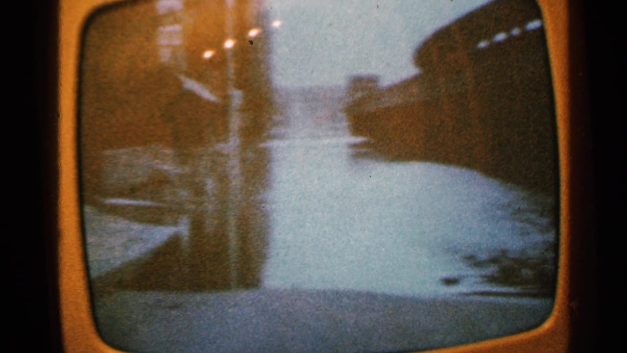 Television screen view of water, shoreline, and dark structures in soft focus.