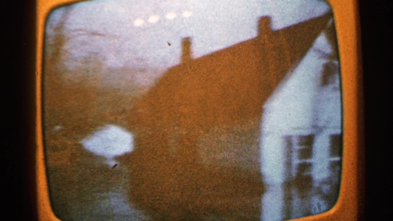 1965 Minnesota 8mm frame of a rounded TV screen showing a house with a steep roof and chimneys, with glare and grain visible.
