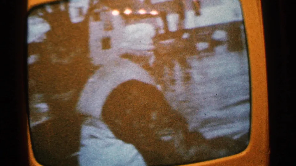 1965 Minnesota home-movie frame with a man in a light shirt near water or boats and a close, partially shadowed face in the foreground.