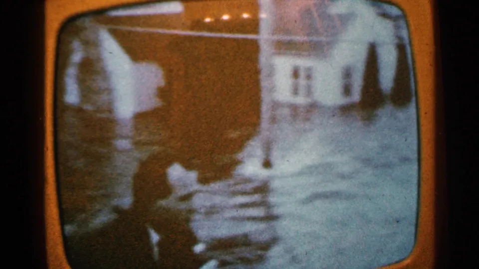 Soft 8mm frame of Minnesota television flood coverage showing pale buildings and waterlogged surroundings.
