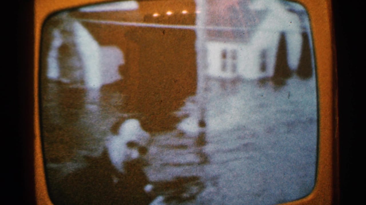 1965 Minnesota home-movie frame seen on a television screen, with a person near floodwater reaching a housefront and steps.