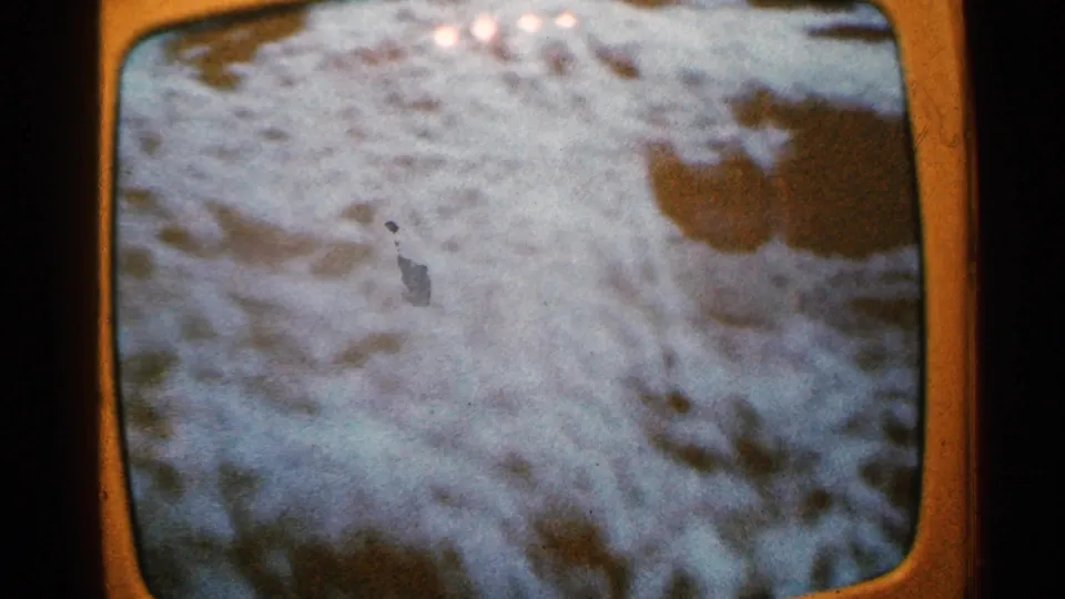 A soft 8mm frame shows turbulent floodwater, with indistinct debris or dark objects visible in the flow.