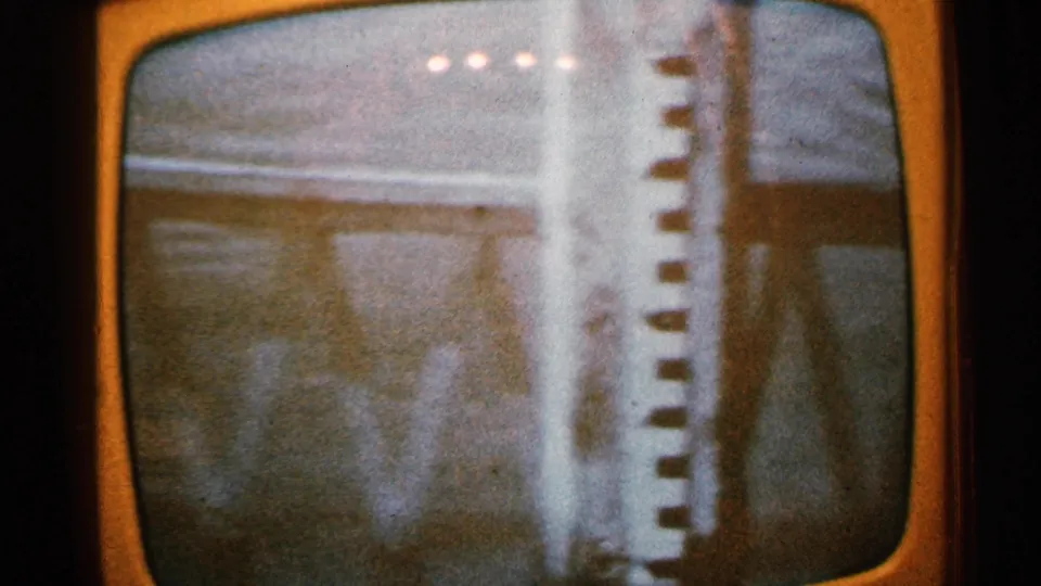 1965 Minnesota flood frame viewed on a television screen, with water near a building edge and a vertical striped object in the foreground.