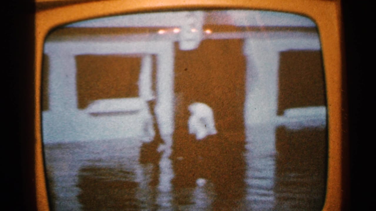 1965 Minnesota home-movie frame showing floodwater against house exteriors, viewed through the curved border of a television screen.